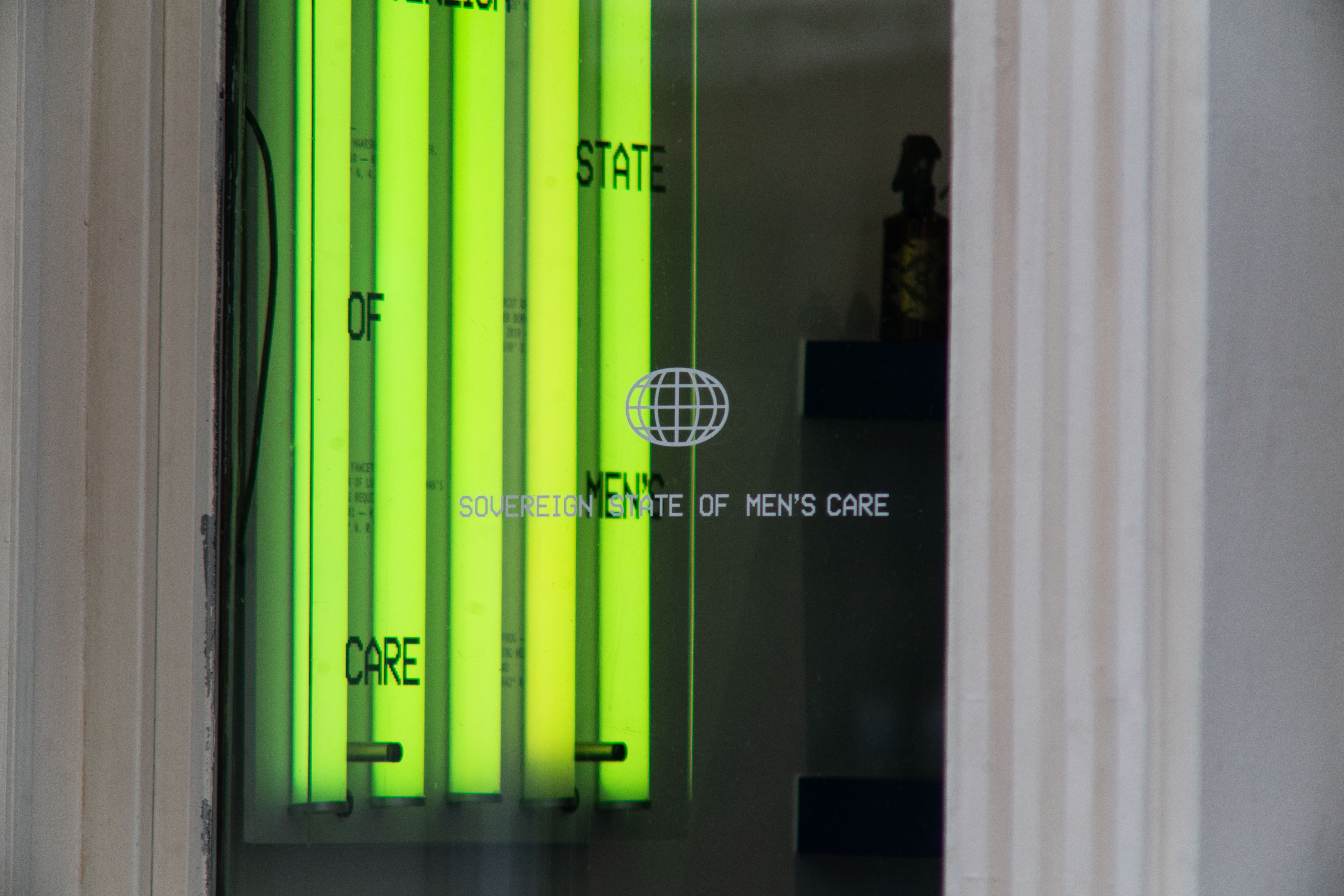 The window from the street — neon tubes behind the etched Sovereign State of Men's Care lettering and globe