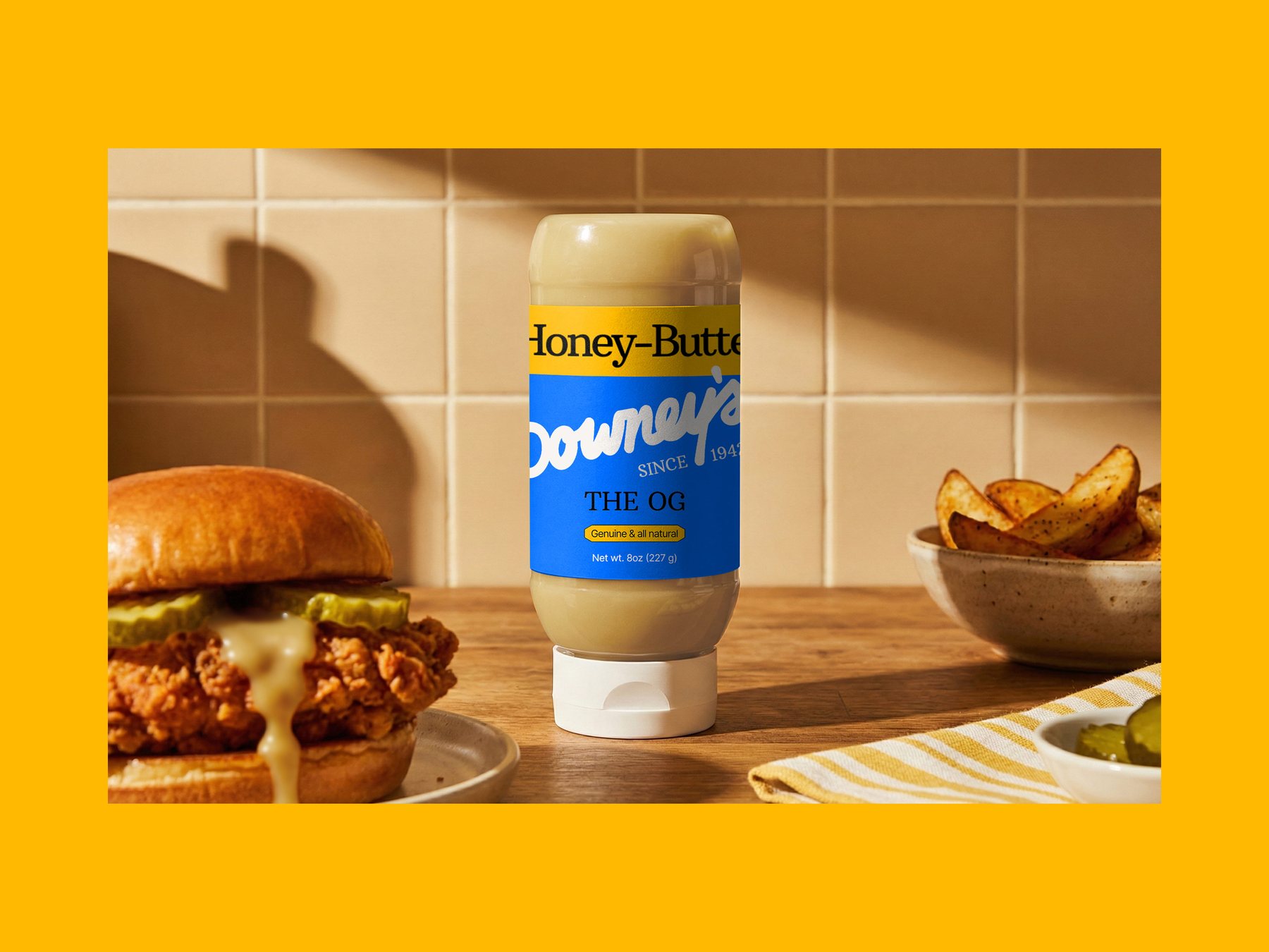 The OG honey butter squeeze bottle on a kitchen counter between a fried chicken sandwich and potato wedges