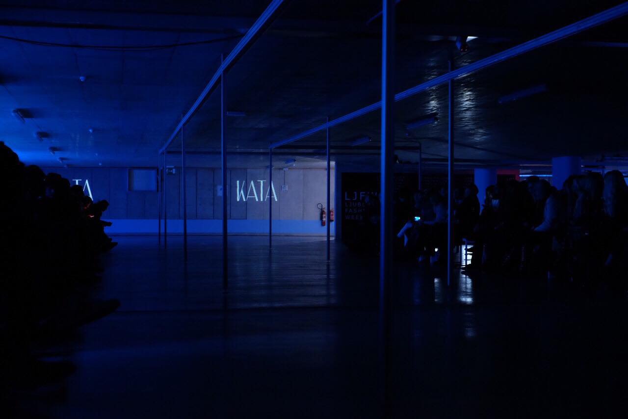 The KATA runway show — a dark blue-lit hall, the wordmark projected on the far wall, the audience waiting in the dark